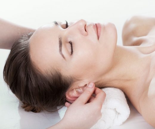 A woman is lying comfortably on her back, receiving an auricular reflexology treatment. A therapist gently presses specific points on her ear using their fingers, focusing on relaxation and therapeutic touch.